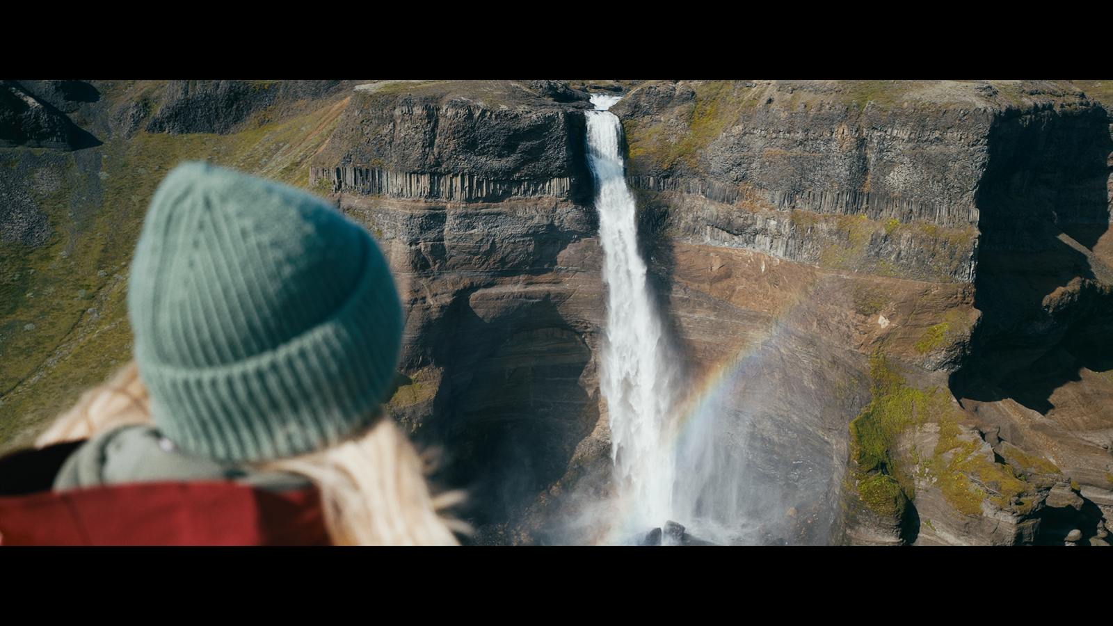 Frau mit Mütze vor einem hohen Wasserfall an einer Felswand